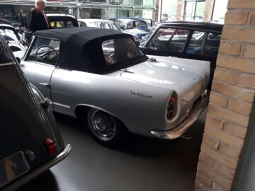 Een zilveren La Mistral-cabriolet die tussen andere voertuigen binnen geparkeerd staat bij Classic Remise, met een persoon vlakbij op de achtergrond. De auto heeft een zwart softtop dak.