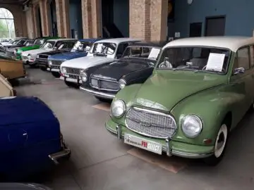 Bij Classic Remise staat binnen op een gladde betonvloer een rij diverse oldtimers geparkeerd. Elke auto heeft een papier met informatie op de voorruit. Op de achtergrond zijn bakstenen muren en grote ramen zichtbaar.