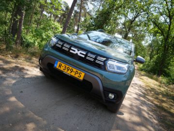 Een groene Dacia Duster met kenteken R-399-PS staat op een zonnige dag geparkeerd op een onverharde weg omgeven door bomen.