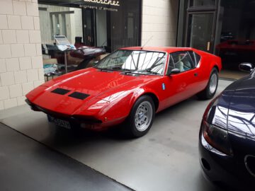 Een rode sportwagen staat geparkeerd in een Classic Remise-showroom met beige tegelwanden en grote glazen ramen.