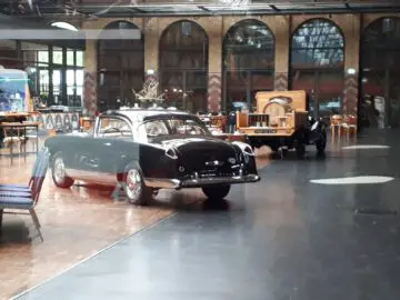 Een oldtimershow in de overdekte showroom van Classic Remise. De ruimte is voorzien van grote boogramen en diverse klassieke voertuigen, waaronder een zwart-witte coupé op de voorgrond.