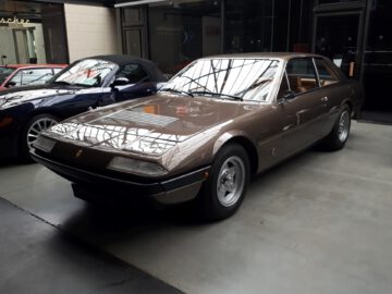 Bij de Classic Remise staat binnen naast andere voertuigen een klassieke bruine sportwagen met een strak design geparkeerd.