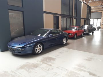 Een rij van drie klassieke auto's, waaronder een blauwe sportwagen, een rode coupé en een zwarte sedan, geparkeerd in de ruime inpandige garage van Classic Remise met moderne scheidingswanden en grote ramen.