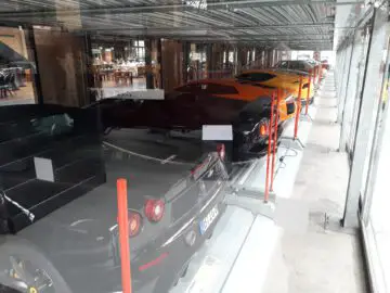 Op de met glas overdekte parkeerplaats van Classic Remise staat een rij sportwagens, waaronder zwarte, oranje en gele modellen.