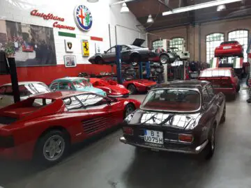 Een goed verlichte autowerkplaats, die doet denken aan Classic Remise, beschikt over meerdere klassieke en sportwagens, waaronder een rode Ferrari die in onderhoud is. Sommige auto's staan op liften, terwijl verschillende autoposters de muren sieren.