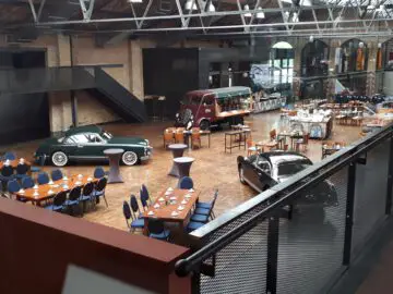 Een overdekte evenementenruimte met oldtimers, gedekte tafels met stoelen en een busje in retrostijl omgebouwd tot bar. De locatie, die doet denken aan Classic Remise, beschikt over hoge plafonds en bakstenen muren die bijdragen aan de tijdloze charme.