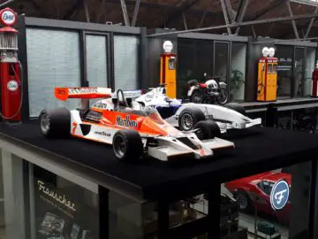 Twee vintage Formule 1-auto's die worden tentoongesteld, één in Marlboro-kleurstelling en de andere in het wit, worden tentoongesteld op een verhoogd platform bij Classic Remise, met oude Esso-benzinepompen en een motorfiets op de achtergrond.