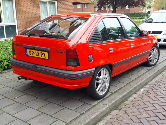 Een rode hatchback Opel Kadett E staat geparkeerd in een woonstraat naast een witte auto, tegen de achtergrond van een bakstenen gebouw en weelderig groen. Op het kenteken staat 