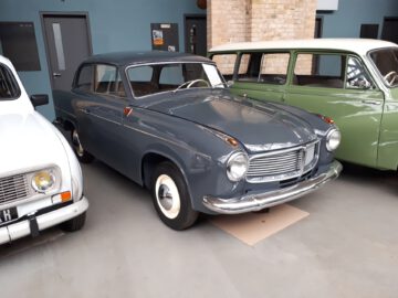 Bij Classic Remise staan drie oldtimers naast elkaar geparkeerd. De centrale auto is een grijze klassieke sedan, geflankeerd door een witte auto aan de linkerkant en een groene aan de rechterkant.