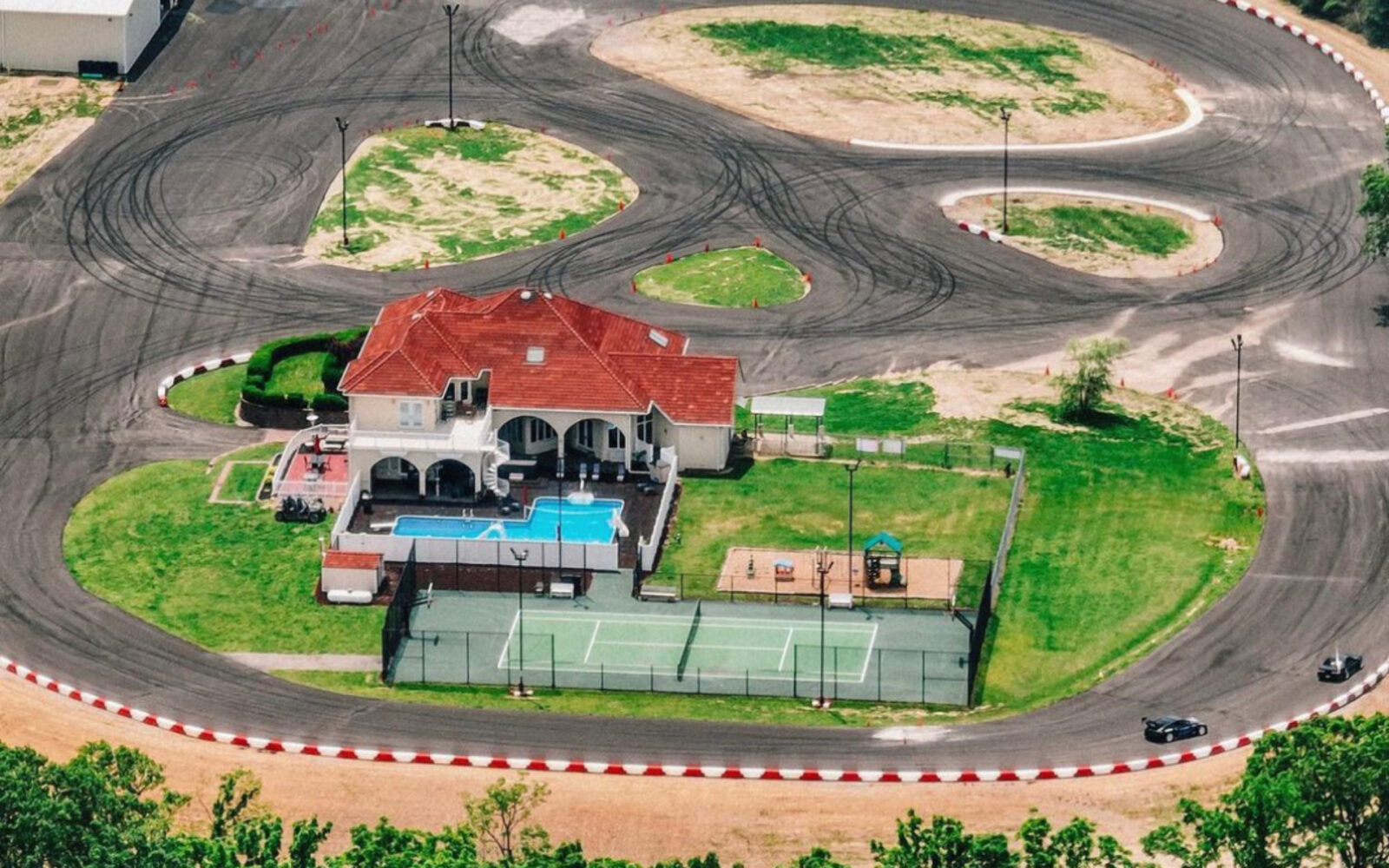 Luchtfoto van een baan met een huis in het midden. Dit drijflandhuis beschikt over een zwembad, een speeltuin en een omheinde tennisbaan, allemaal omgeven door het racecircuit.