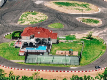 Luchtfoto van een baan met een huis in het midden. Dit drijflandhuis beschikt over een zwembad, een speeltuin en een omheinde tennisbaan, allemaal omgeven door het racecircuit.