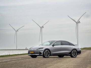 Een zilveren Hyundai staat geparkeerd op een weg in de buurt van een watermassa, met drie grote windturbines op de achtergrond onder een bewolkte hemel.
