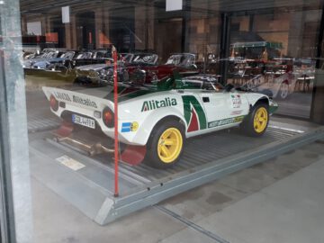 Een witte en groene Lancia Stratos-rallyauto met Alitalia-kleurstelling en gele wielen wordt tentoongesteld in de Classic Remise-showroom, omringd door andere vintage klassiekers.