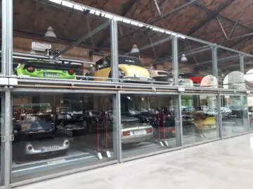 Een indoor autodisplay met meerdere niveaus in de Classic Remise toont vintage en moderne voertuigen, waaronder een groene, gele en rode auto op de bovenste verdieping en verschillende andere daaronder, allemaal in een met glas omsloten structuur.