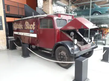 Een vintage rode bestelwagen met 'Wittler Brot'-opschrift op de zijkant wordt prominent getoond in de Classic Remise. De motorkap is open waardoor de motor zichtbaar is, terwijl andere voertuigen gedeeltelijk zichtbaar zijn op de achtergrond.