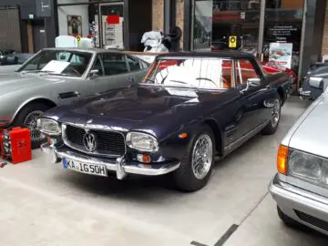 In de overdekte showroom van Classic Remise staat een klassieke blauwe Maserati-auto geparkeerd, naast andere oldtimers.