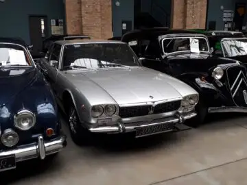 Bij Classic Remise staat een zilveren vintage auto geparkeerd, tussen twee andere klassieke auto's. Op de grille aan de voorzijde van de auto staat een drietandsymbool.