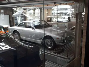 Een zilveren klassieke sportwagen wordt tentoongesteld in een kamer met glazen wanden in een garage bij Classic Remise, waarbij de zijkant en een deel van de voorkant zichtbaar zijn.