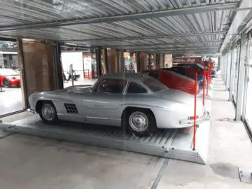 Twee klassieke auto's, waaronder een zilveren Mercedes-Benz 300SL en een rode Ferrari, staan geparkeerd op een autoopslagplatform met meerdere niveaus in de beroemde Classic Remise-garage.