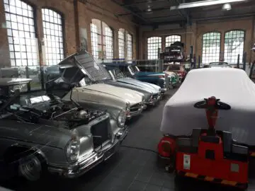 Een rij klassieke auto's wordt tentoongesteld in de showroom met bakstenen muren van Classic Remise; sommige hebben open kappen. Op de voorgrond staat een gedeeltelijk overdekte auto op een rode wagen.