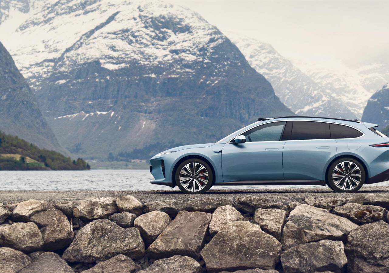 Een blauwe Nio ET5-toerwagen staat geparkeerd op een rotsachtige ondergrond met op de achtergrond besneeuwde bergen en een meer.