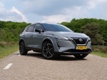 Een zilveren Nissan Qashqai e-Power SUV staat geparkeerd op een verharde weg, omgeven door groen op een heldere dag. Op het kenteken staat "R-044-JV.