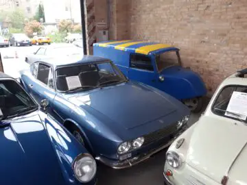 Een verzameling oldtimers in een garage, met onder meer een blauwe sportwagen en een blauw-gele bestelwagen, die doet denken aan de klassieke elegantie die je bij Classic Remise aantreft.