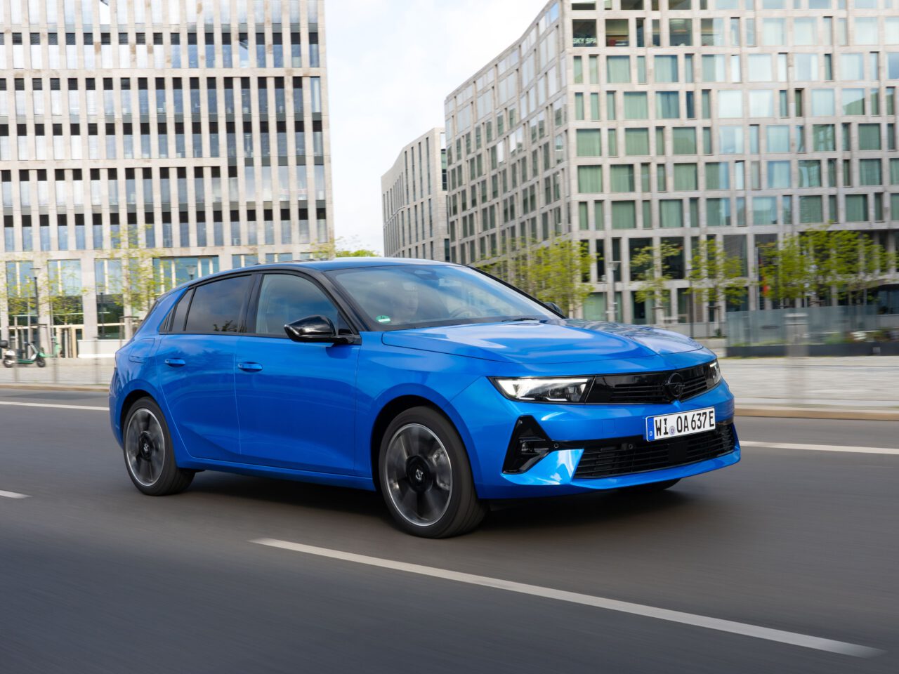Een blauwe Opel Astra Electric hatchback rijdt door een stadsstraat met moderne glazen gebouwen op de achtergrond.
