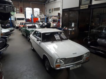 De Classic Remise herbergt een verzameling oldtimers die binnenshuis worden tentoongesteld, met een witte Peugeot 304 prominent op de voorgrond en daarachter staan verschillende andere klassieke auto's opgesteld.