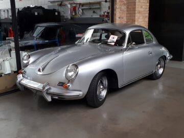 Een klassieke zilveren coupé staat binnen geparkeerd op een betonnen vloer met een bakstenen muur en een glazen raam op de achtergrond, wat doet denken aan de retrocharme van Classic Remise. Op de voorruit wordt een bord geplaatst.