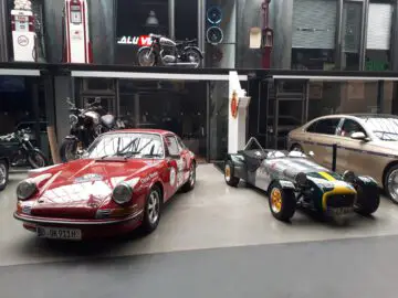 Een rode vintage Porsche en een groene Lotus Seven staan geparkeerd in Classic Remise naast een motorfiets. Op de achtergrond zijn extra auto's en voertuigonderdelen zichtbaar, wat bijdraagt aan de nostalgische sfeer van het automuseum.