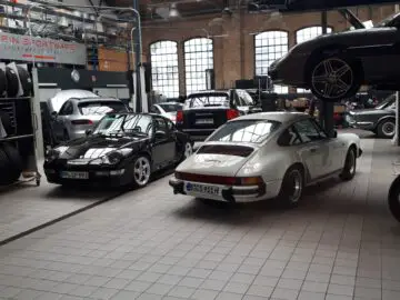 Diverse sportwagens, waaronder zwart-witte Porsche-modellen, staan in een ruime garage met hoge plafonds en grote ramen bij Classic Remise. Op de achtergrond zijn bandenrekken, autoliften en gereedschappen zichtbaar.