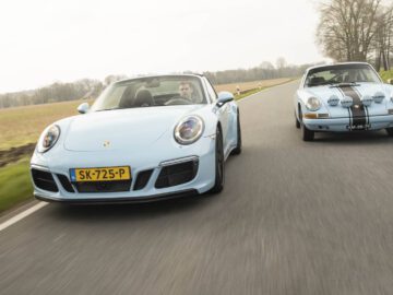 Twee lichtblauwe Porsche-sportwagens die sierlijk over een plattelandsweg navigeren. De auto vooraan heeft het kenteken SK-725-P, terwijl de auto erachter AM-97-79 draagt. Velden en bomen vormen het idyllische decor.