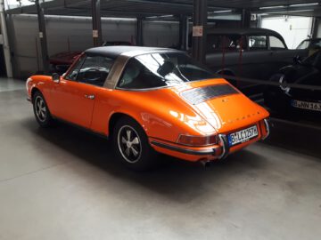 Een vintage oranje Porsche 911 Targa staat binnen geparkeerd bij Classic Remise en toont zijn tijdloze ontwerp en de achterste kentekenplaat met de tekst "B-UC 9125H.