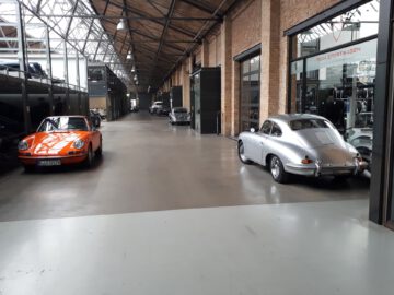 Een ruime inpandige garage, die doet denken aan Classic Remise, beschikt over klassieke auto's, waaronder een oranje en een zilverkleurig voertuig, geparkeerd langs bakstenen muren en grote glazen ramen.