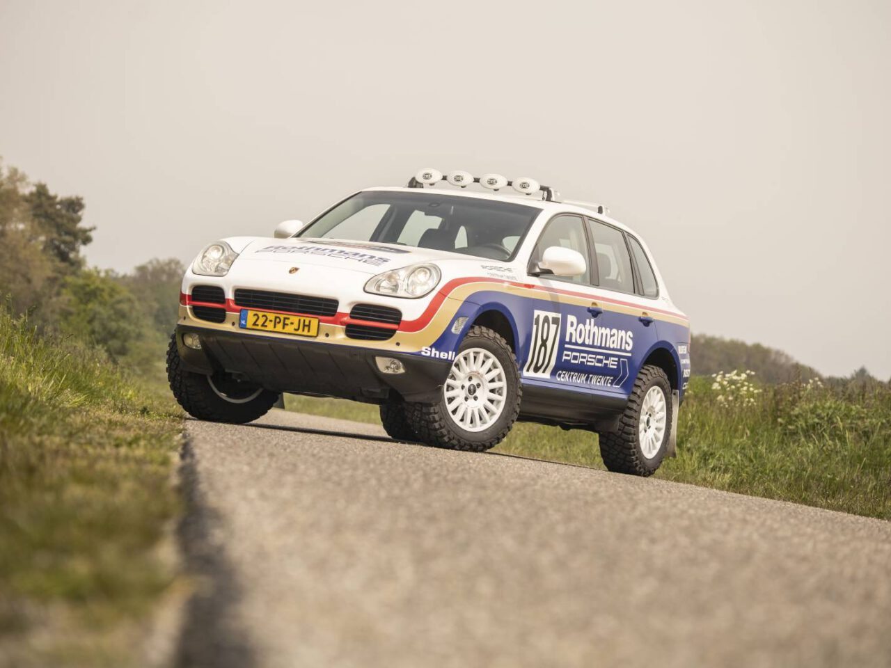 Een gemodificeerde Porsche Cayenne met een Rothmans-kleurstelling en een geest die doet denken aan de Porsche Dakar, geparkeerd op een verharde weg, uitgerust met offroad-banden en extra verlichting op het dak.