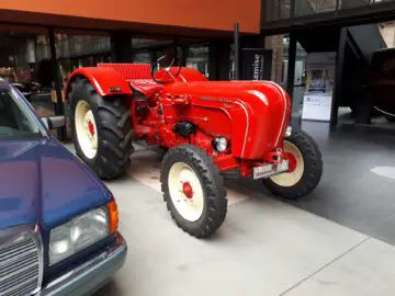 Een rode tractor met grote banden wordt naast een blauw voertuig tentoongesteld in een binnenomgeving geïnspireerd op het moderne industriële ontwerp van Classic Remise.