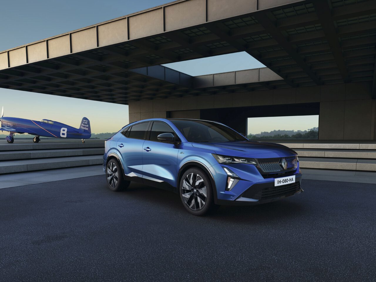 Een blauwe Renault Rafale SUV staat geparkeerd op een moderne oprit met een blauw vintage vliegtuig aan de linkerkant en een eigentijds gebouw op de achtergrond, alles onder een heldere hemel.