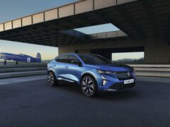 Een blauwe Renault Rafale SUV staat geparkeerd op een moderne oprit met een blauw vintage vliegtuig aan de linkerkant en een eigentijds gebouw op de achtergrond, alles onder een heldere hemel.