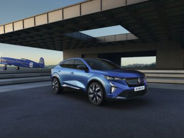 Een blauwe Renault Rafale SUV staat geparkeerd op een moderne oprit met een blauw vintage vliegtuig aan de linkerkant en een eigentijds gebouw op de achtergrond, alles onder een heldere hemel.