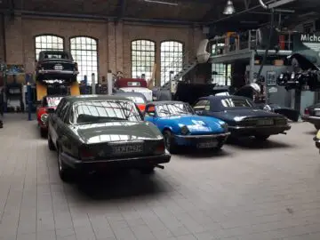 Een garage met meerdere oldtimers, waaronder een groene sedan, een blauwe cabriolet en een zwarte auto, allemaal geparkeerd op een tegelvloer die doet denken aan Classic Remise, met gereedschap en uitrusting zichtbaar op de achtergrond.