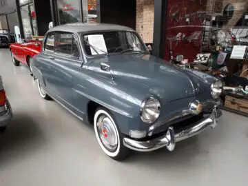 Een vintage grijze sedan uit het midden van de 20e eeuw, tentoongesteld in Classic Remise, een overdekte autotentoonstelling, omringd door memorabilia van klassieke auto's.