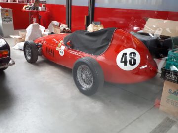 Een vintage rode raceauto met het nummer 48 op de zijkant, binnen geparkeerd bij Classic Remise naast verschillende dozen en auto-items.