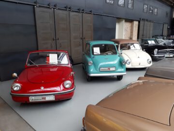 Een rij vintage brommobielen tentoongesteld in de Classic Remise-showroom, waaronder een rode auto, een lichtblauwe auto en een beige auto, met grotere klassieke auto's op de achtergrond.