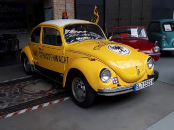 Een gele oldtimer met "ADAC-Strassenwacht" op de zijkant, binnen geparkeerd bij Classic Remise naast andere klassieke auto's. De auto is voorzien van een verlichting aan de bovenkant en historische kentekenplaten, waardoor zijn charme in een museumomgeving wordt getoond.