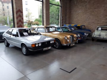 Bij Classic Remise staat een rij oldtimers, waaronder een zilveren hatchback, een gouden sedan en een blauwe cabriolet, binnen geparkeerd op een gepolijste betonnen vloer tegen een bakstenen muur met grote ramen.