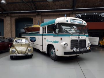 Een oldtimer-autotransporter met een boot erop staat geparkeerd naast een klassieke gele Volkswagen Kever in Classic Remise, waardoor in het magazijn een auto-tijdcapsule ontstaat.