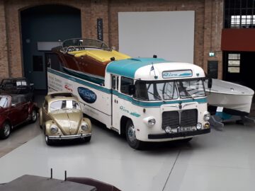 Een vintage boottransportwagen met een houten boot staat binnen geparkeerd. Ernaast staat een klassieke auto. De scène, onderdeel van het Classic Remise museum of de tentoonstelling, straalt tijdloze elegantie en autogeschiedenis uit.