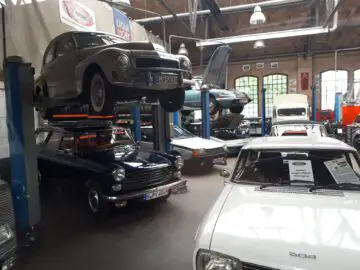 Een autowerkplaats bij Classic Remise toont verschillende klassieke auto's, waaronder een zilveren auto op een lift, een zwarte stationwagen, een witte sedan en een blauwe cabriolet met open kap. De ruimte is voorzien van grote ramen en bovenlichten.