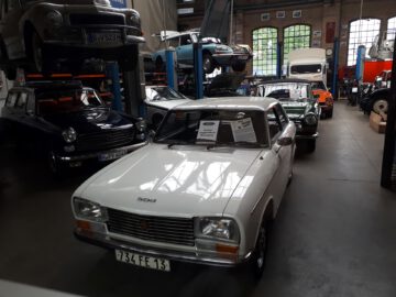 Een verzameling klassieke auto's tentoongesteld in een garage van de Classic Remise, met een witte 504 op de voorgrond en diverse andere oldtimers, waaronder een groene 124 Spider en een oranje 2002, die de achtergrond sieren.